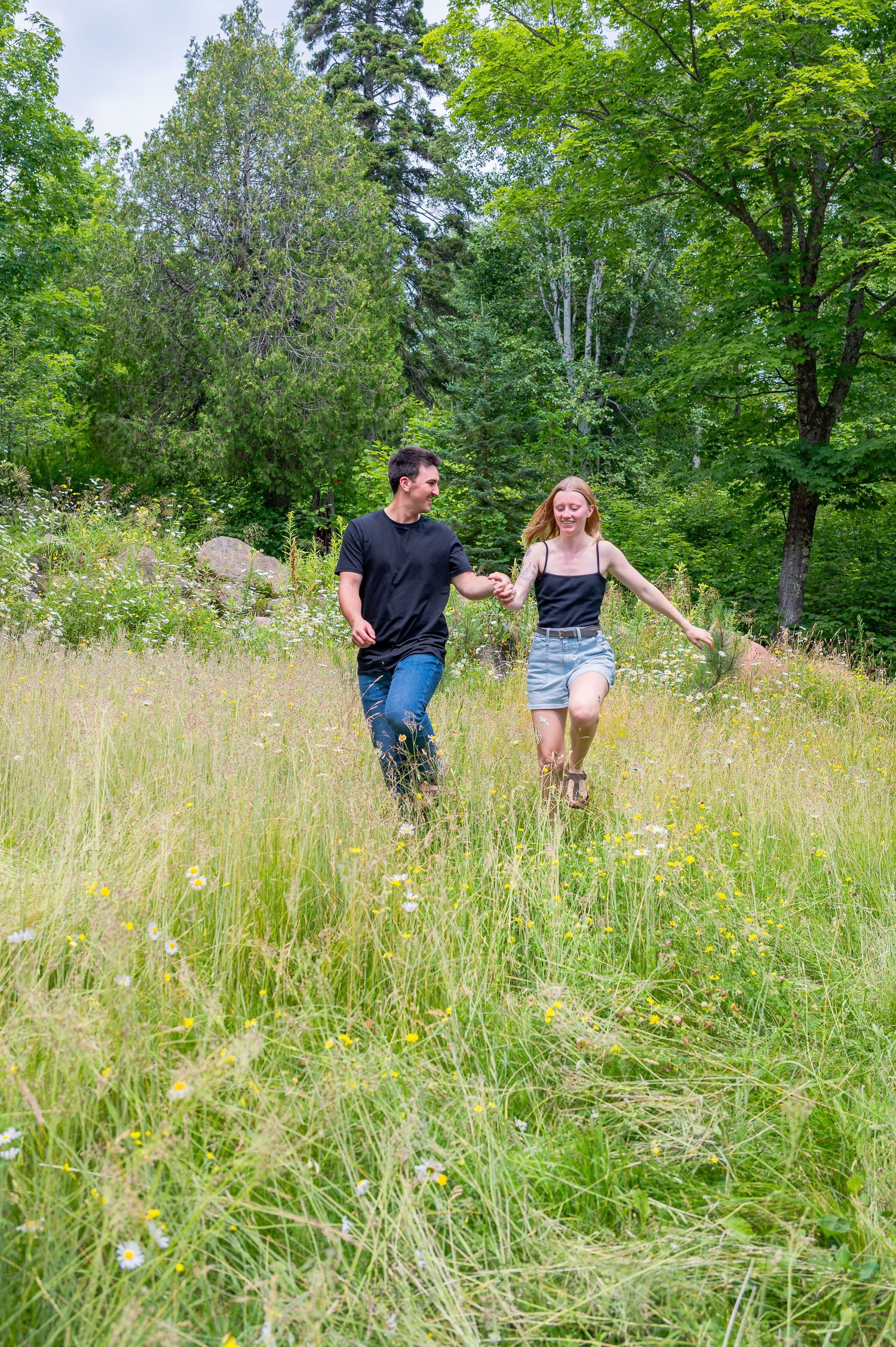 A couple walks hand in hand through a sunny meadow with tall grass and trees in the background.