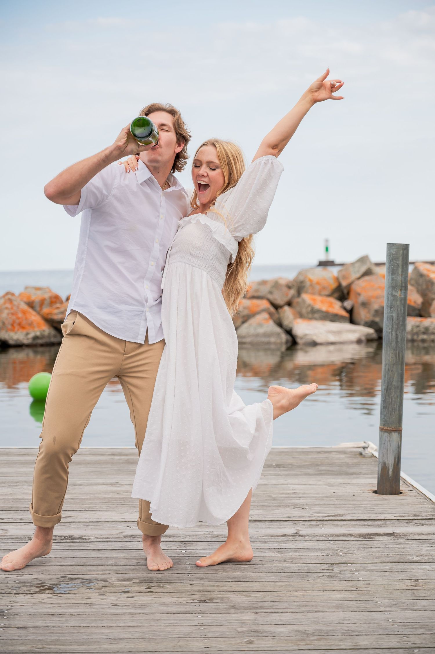 Two people in casual attire dancing and celebrating on a wooden dock by the water with rocks in the background.