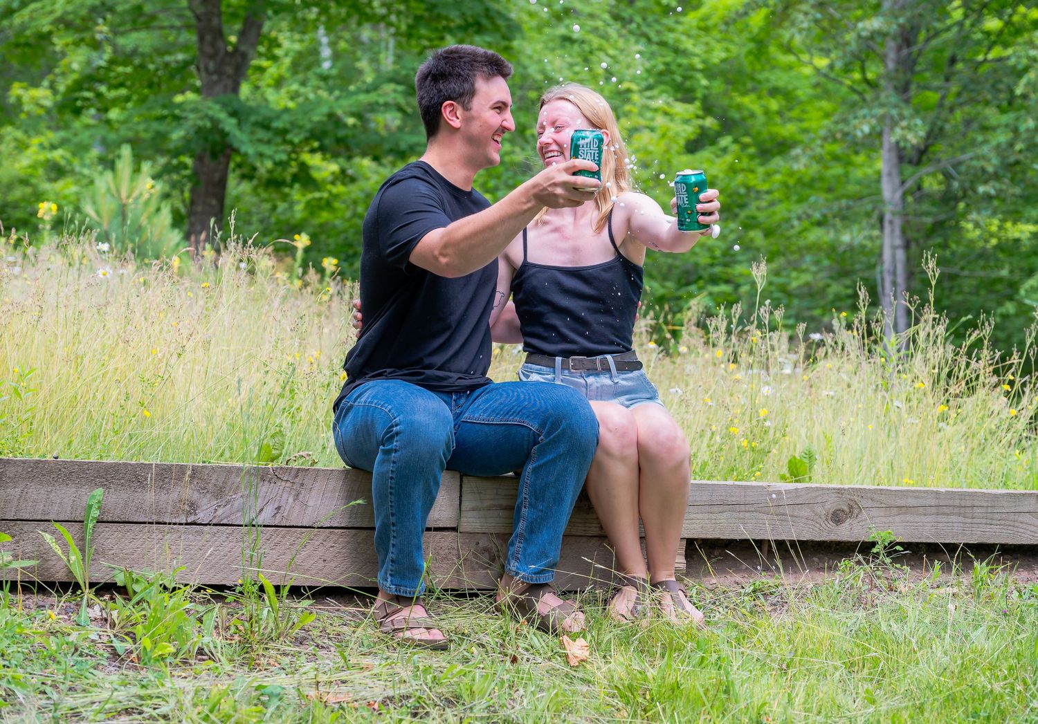 Two people sit casually on a wooden bench in a lush summer meadow sharing drinks together.