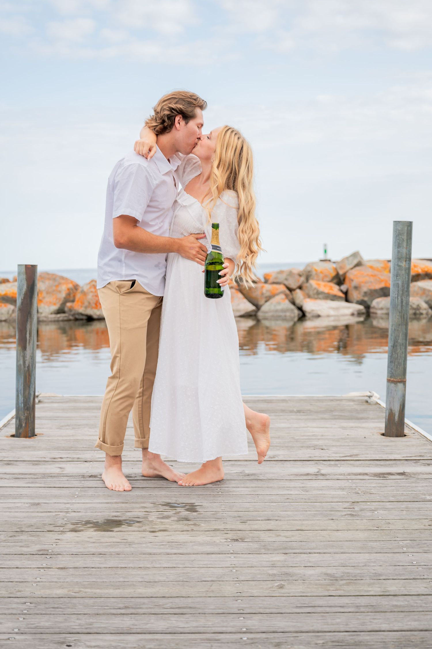 A romantic couple shares a kiss on a wooden pier while holding a beverage bottle against a seaside backdrop of rocks.