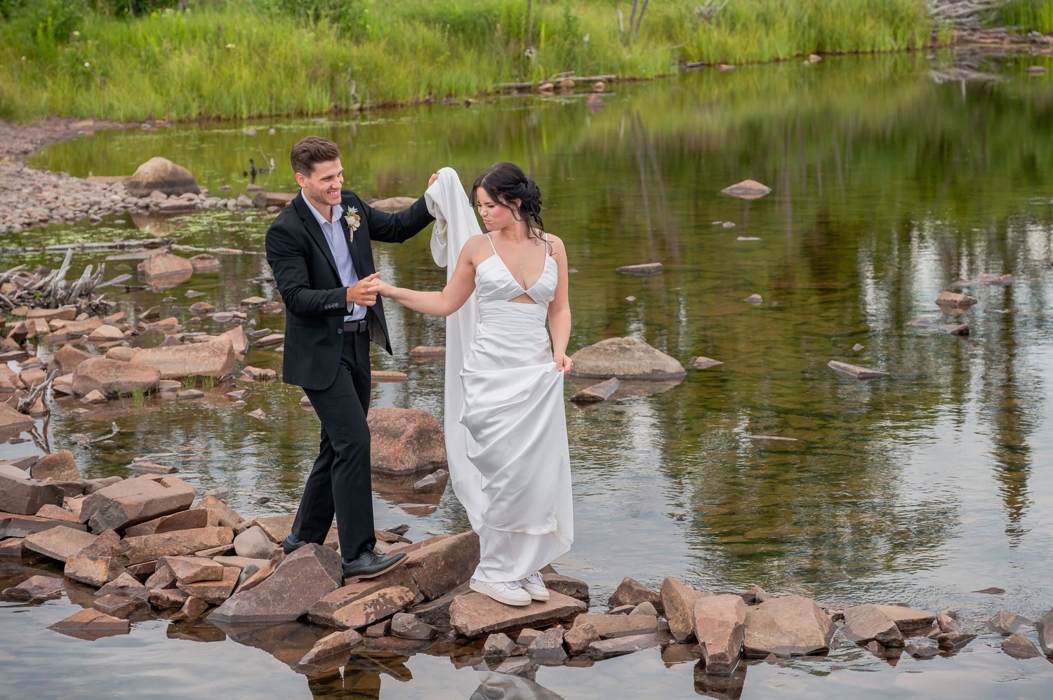 A couple celebrates their wedding day in a scenic lakeside setting with stone outcroppings near the water's edge.