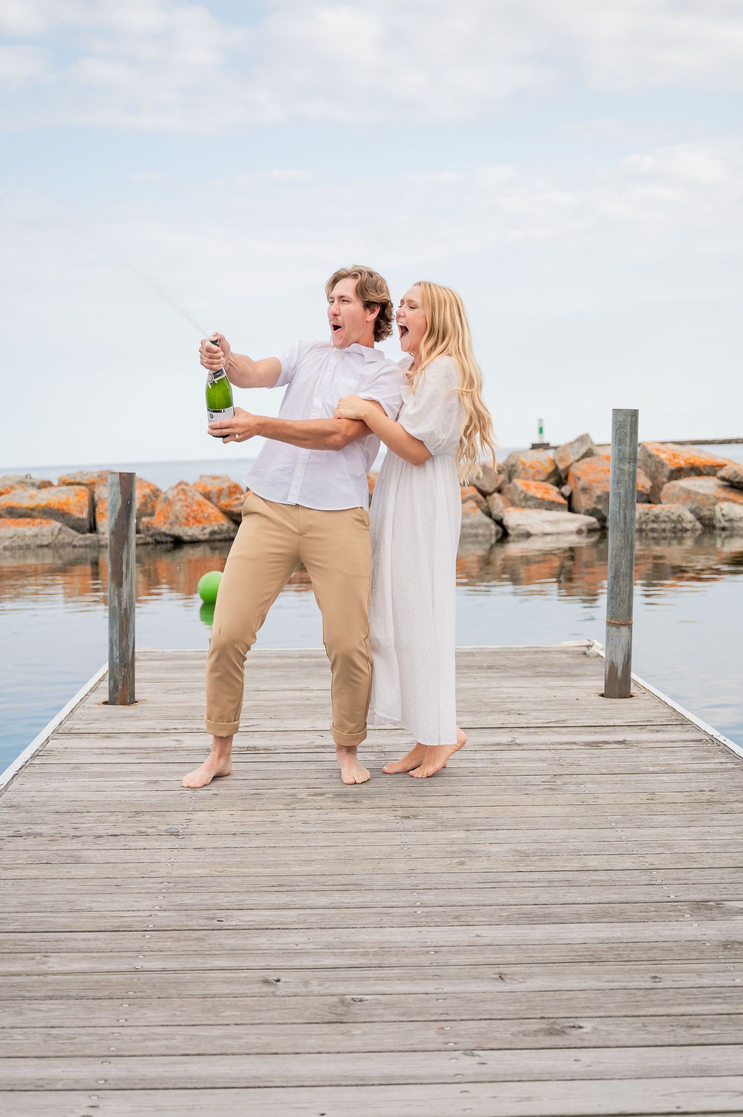 Two people in casual attire celebrating with champagne on a wooden dock by the ocean at sunset.