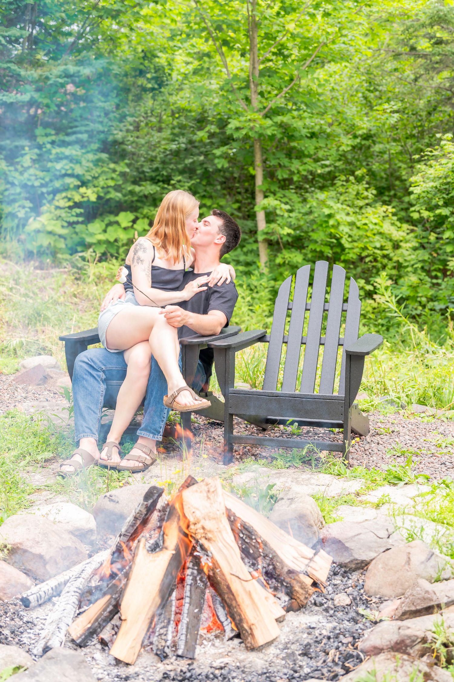 A couple sits together on an Adirondack chair beside a glowing campfire in a green forest setting.