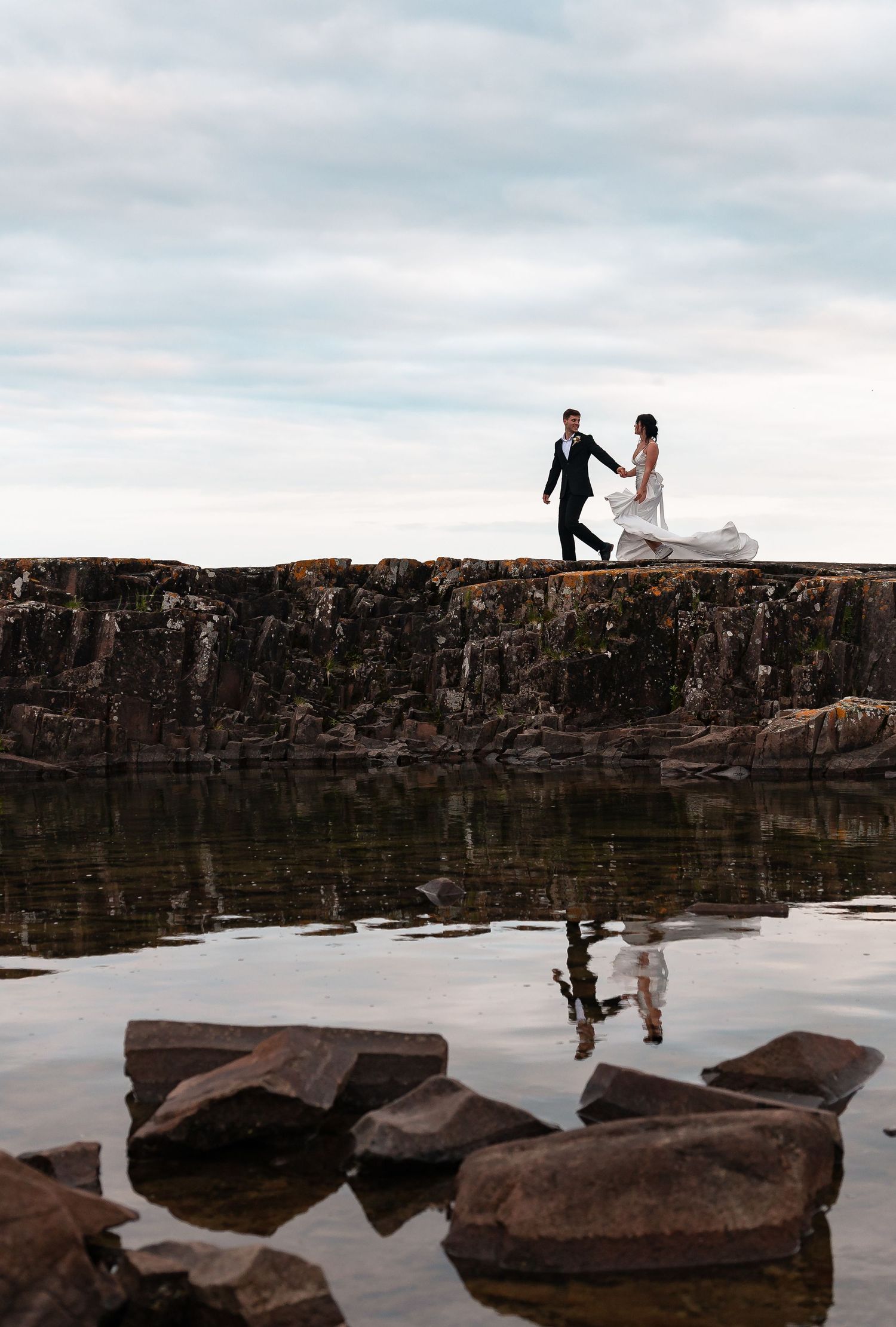 Two silhouettes stand on a rocky cliff edge overlooking tranquil water with their reflections mirrored in the still surface below.