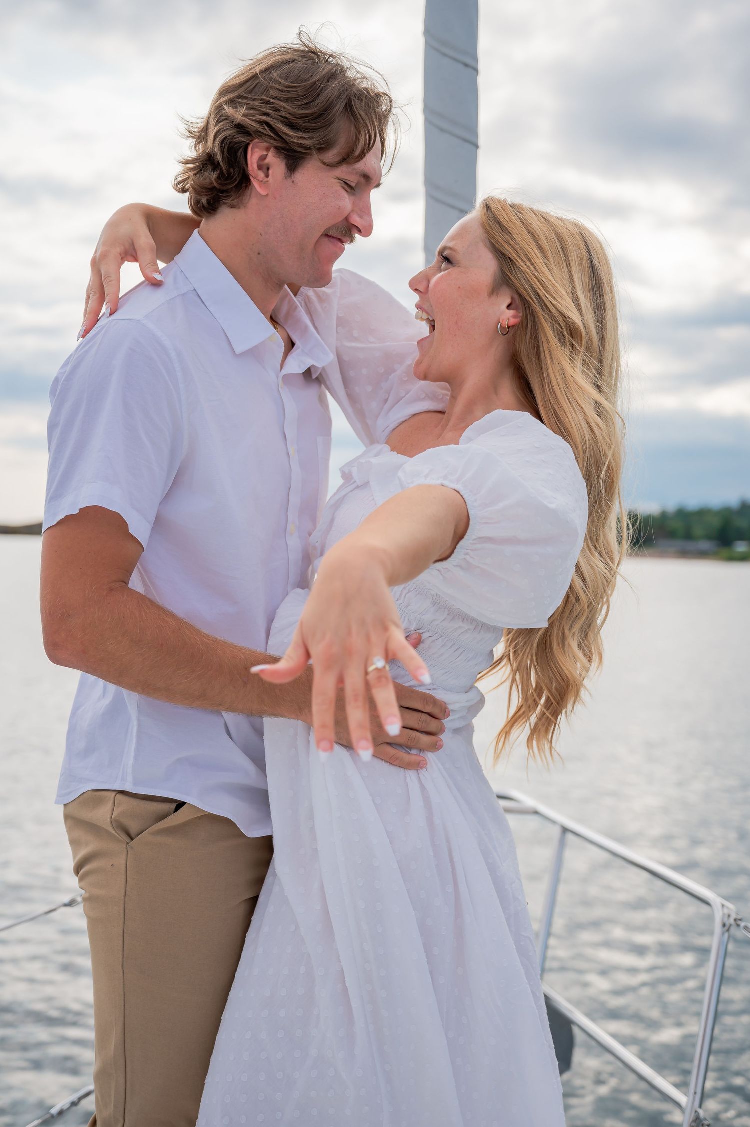 A couple in white clothing shares a romantic moment while standing on a boat deck with water and cloudy skies in the background.