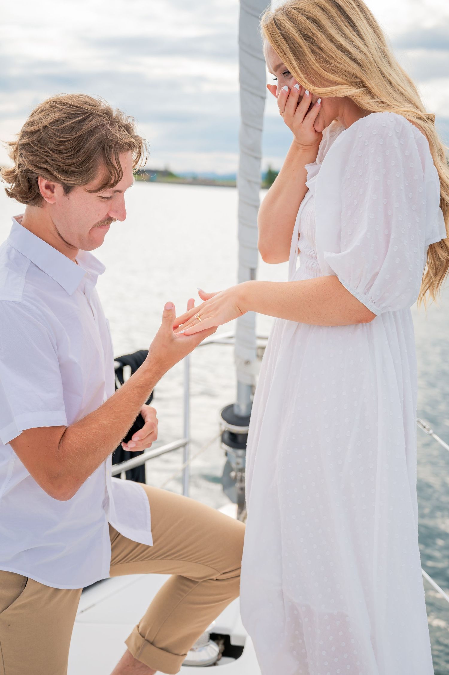 A couple shares a romantic proposal moment on a boat deck with water and cloudy skies in the background.