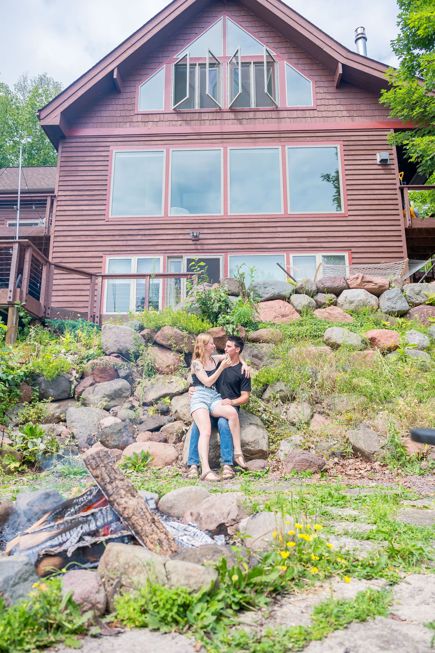 A rustic brown cabin with large windows sits on a rocky hillside surrounded by green vegetation and landscaping.