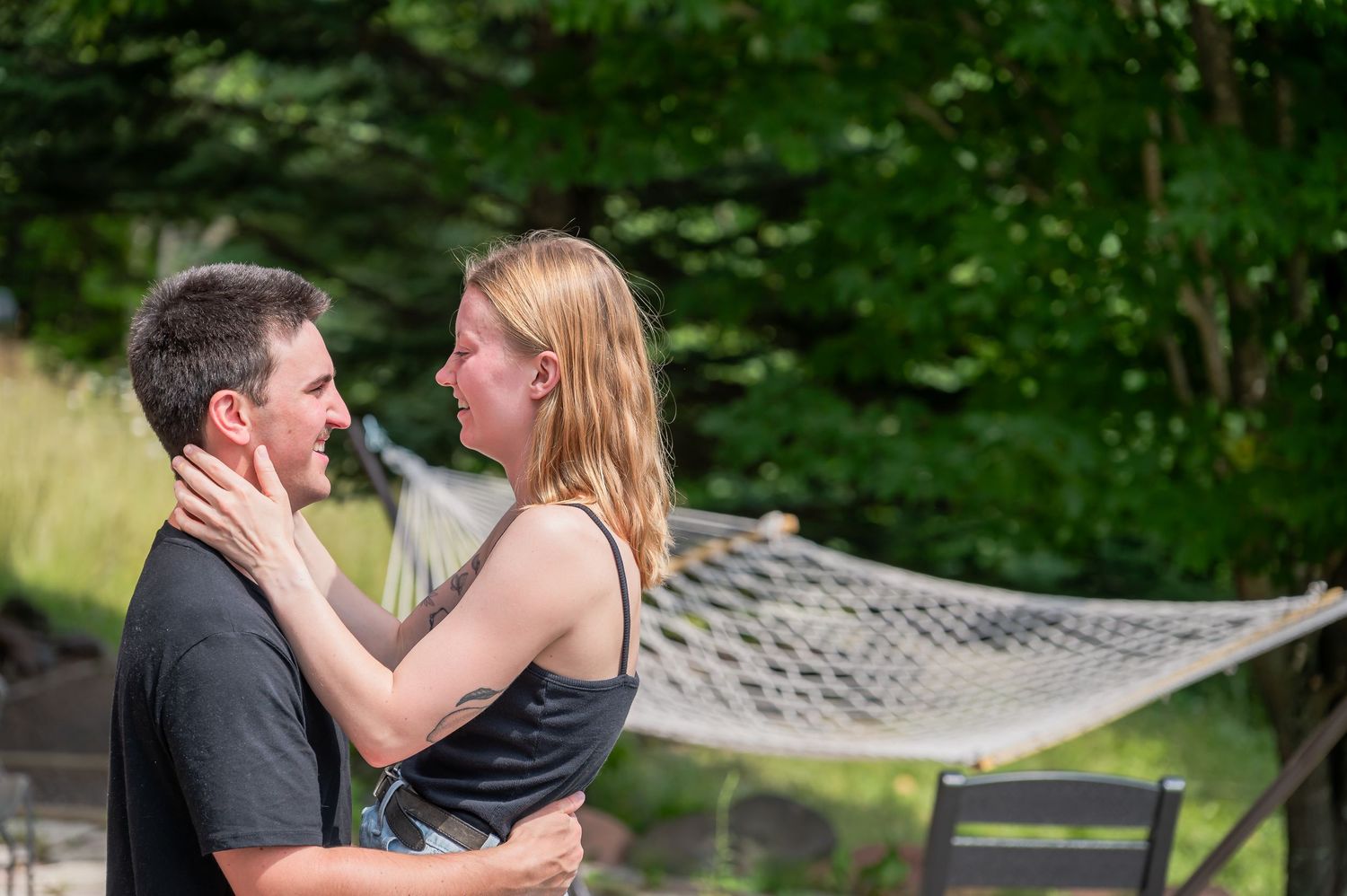 A romantic couple embraces intimately outdoors near a hammock with lush greenery in the background.