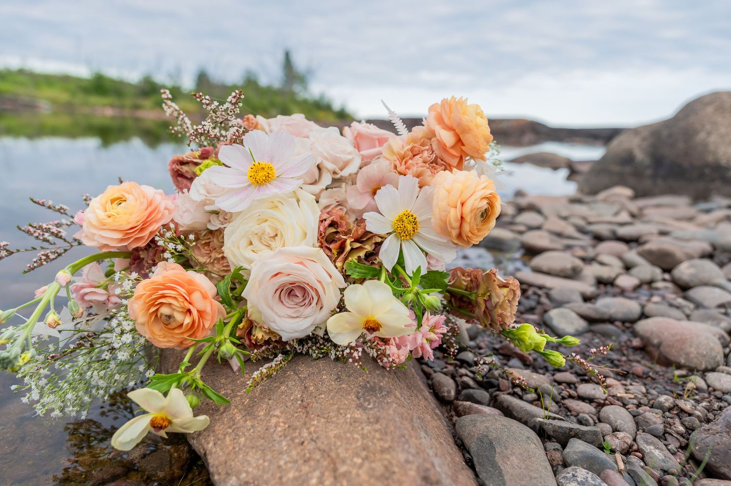 Peach and cream floral bouquet with roses and daisies arranged on rocky riverbank against scenic water backdrop.