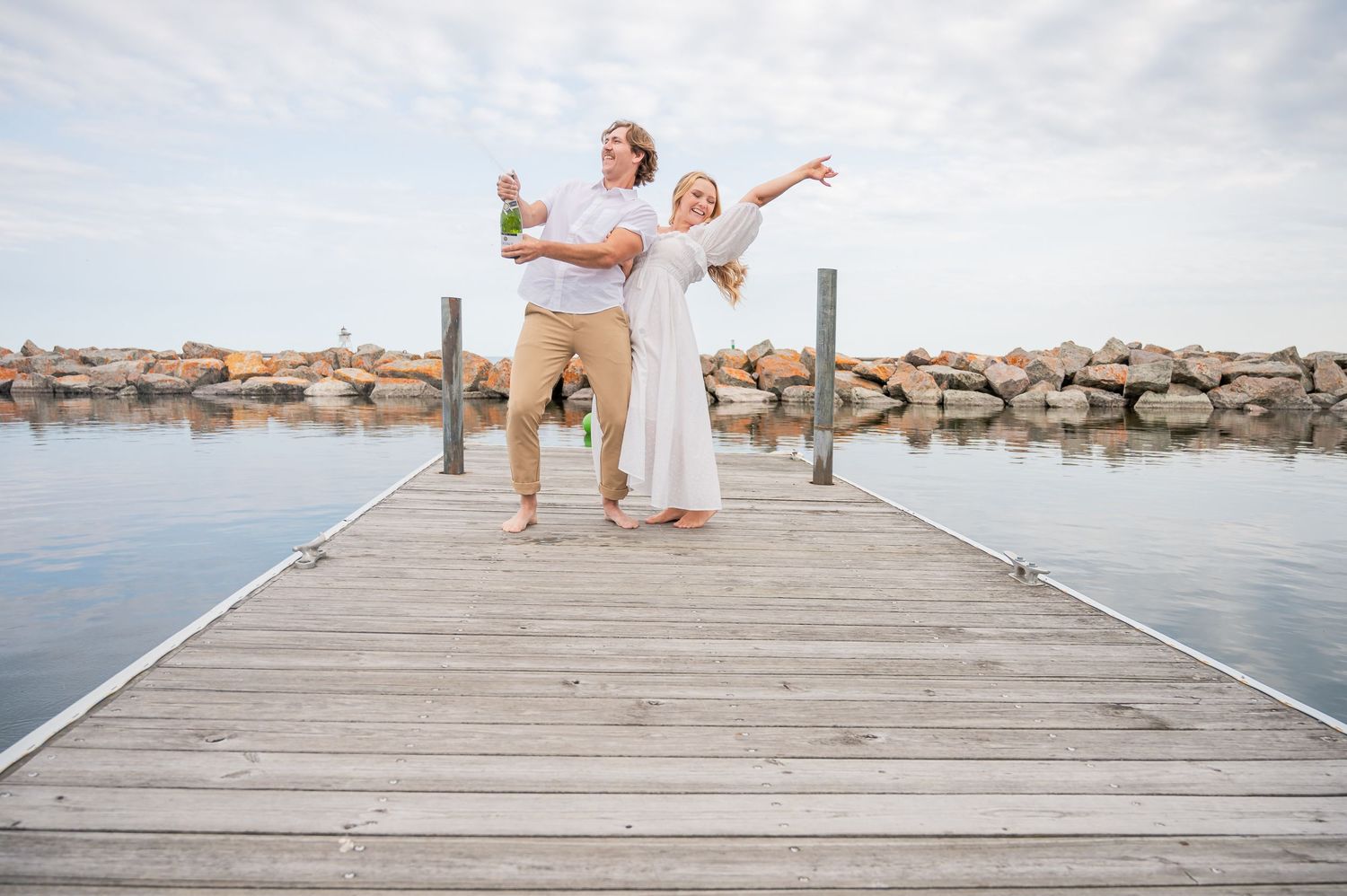 A couple in white attire strikes joyful poses on a wooden pier overlooking a rocky shoreline and calm waters.