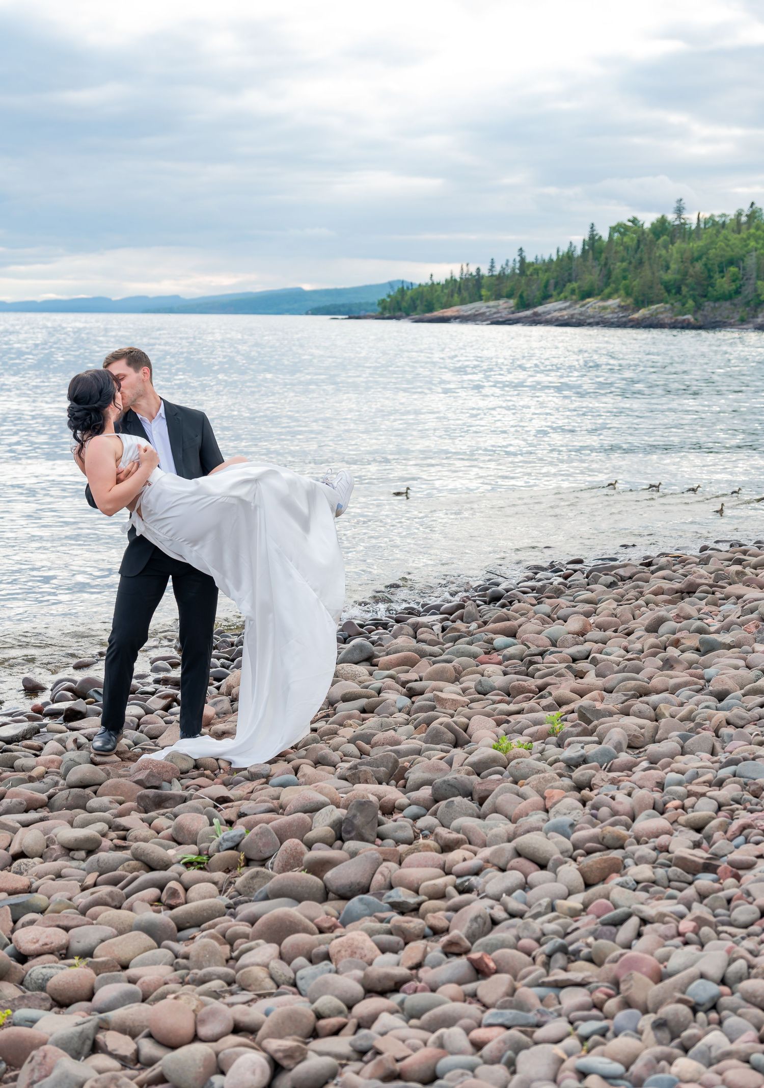 A romantic lakeside moment on a rocky beach with mountains and forest in the distance.