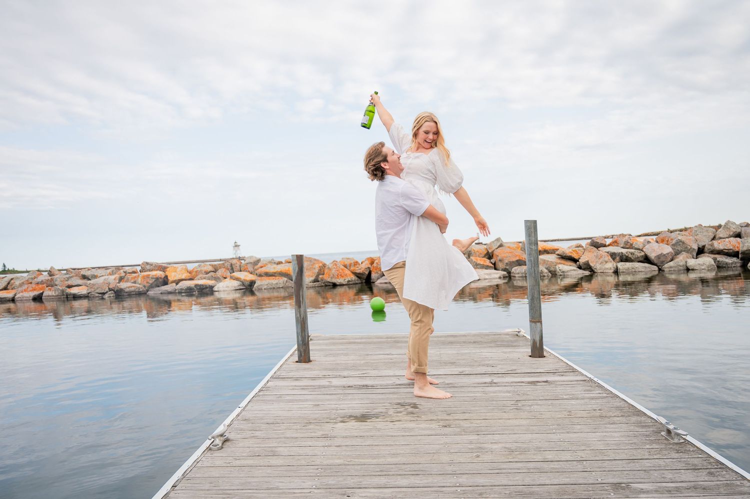 A couple in white clothing celebrates joyfully on a wooden pier overlooking calm waters.