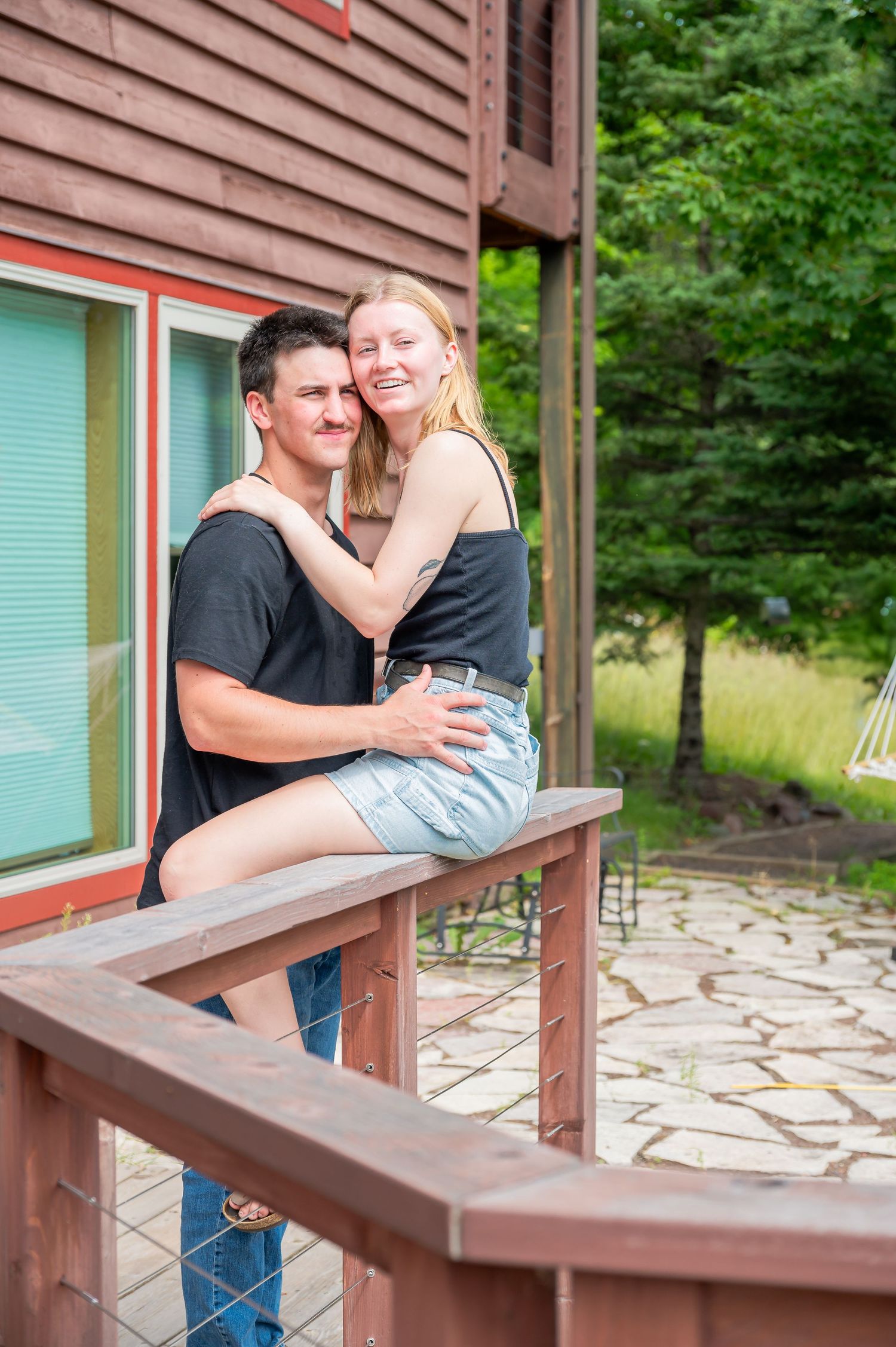 A couple embraces on a wooden deck outside a brown house with turquoise trim and stone patio on a sunny summer day.