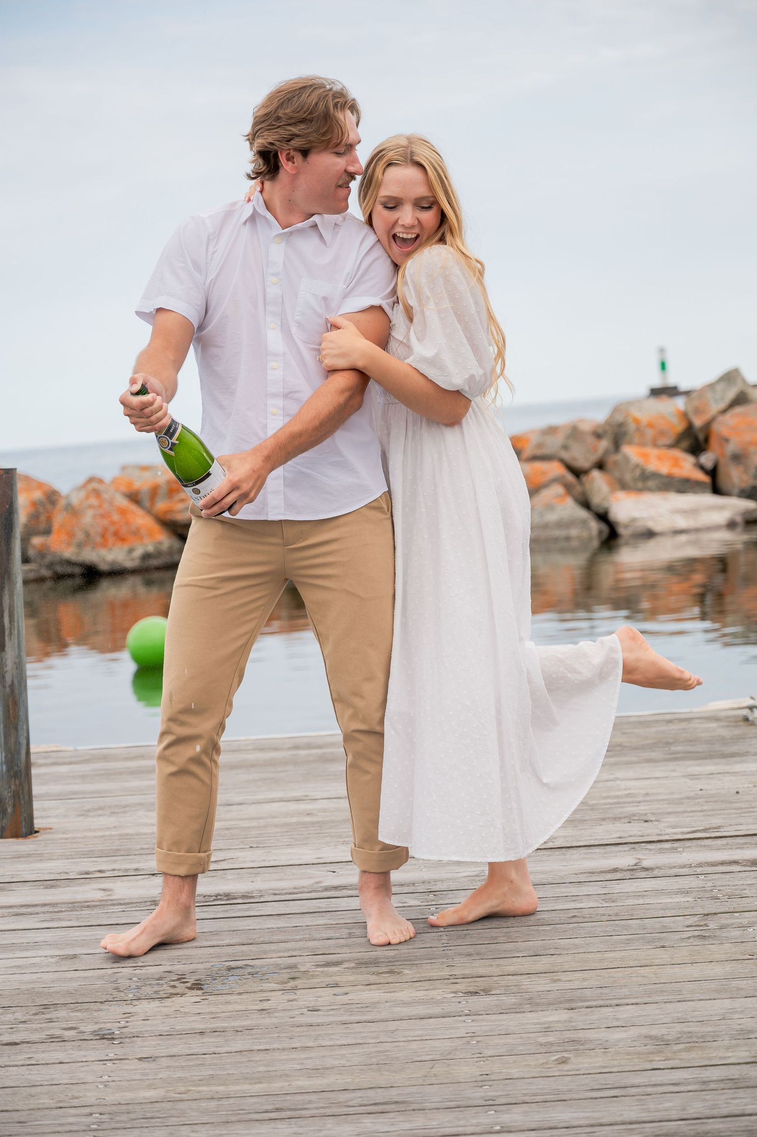 A couple celebrates joyfully on a wooden dock near rocky shore, with person holding champagne bottle.