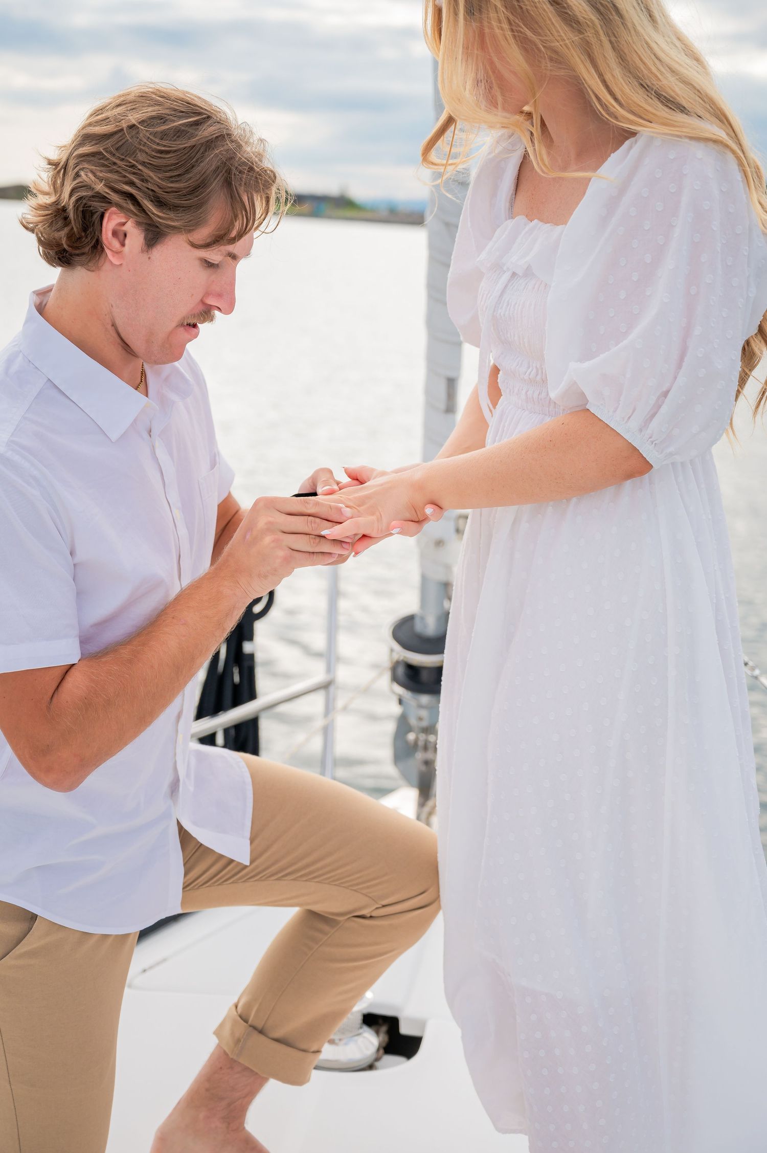 A romantic proposal moment on a boat dock with someone in a white dress and someone kneeling in khakis and white shirt.