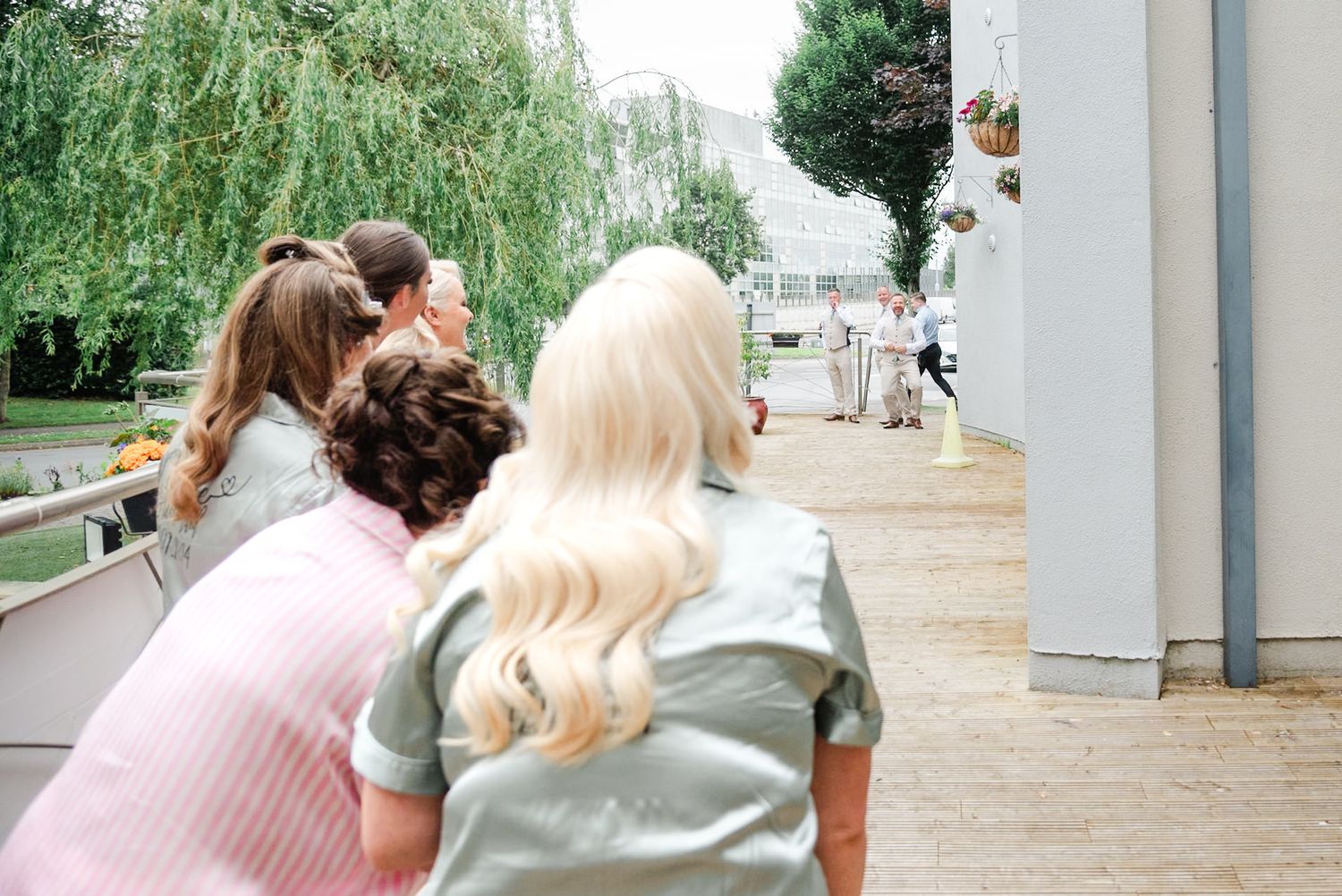 A group watches an attraction through a fence outside a building at a park or venue with willow trees in the background.