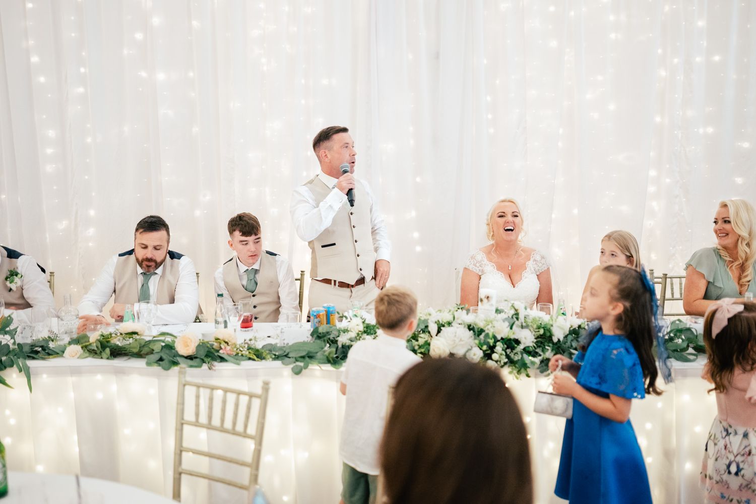Wedding party stands at a decorated head table with lush greenery garlands and elegant white decor during reception.