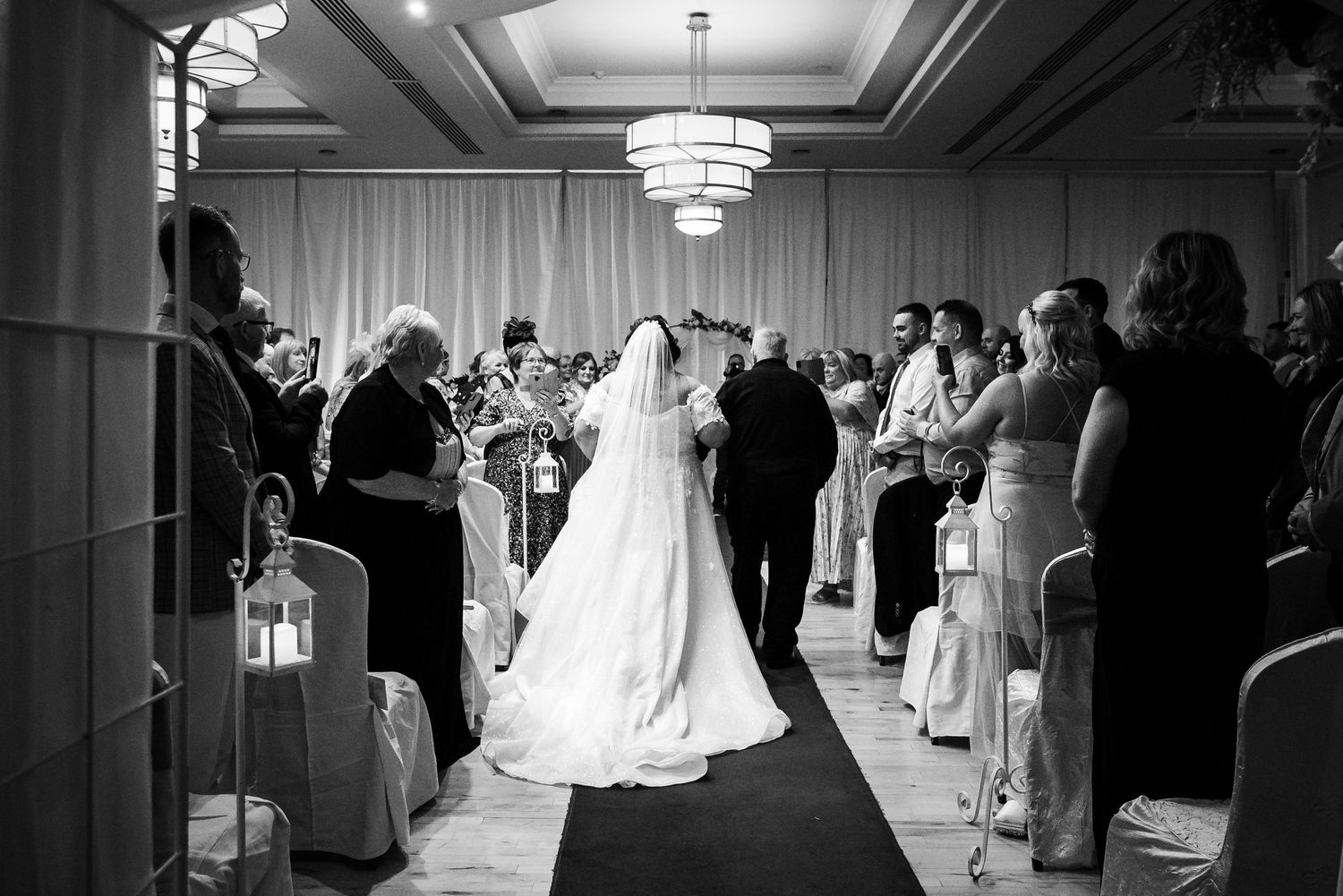 Black and white photograph of a wedding ceremony in an elegant ballroom with crystal chandeliers and guests lining the aisle.