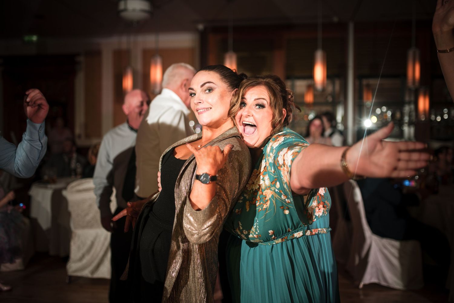Two wedding guests in elegant dresses having fun singing and dancing at a reception party with dimmed lighting.