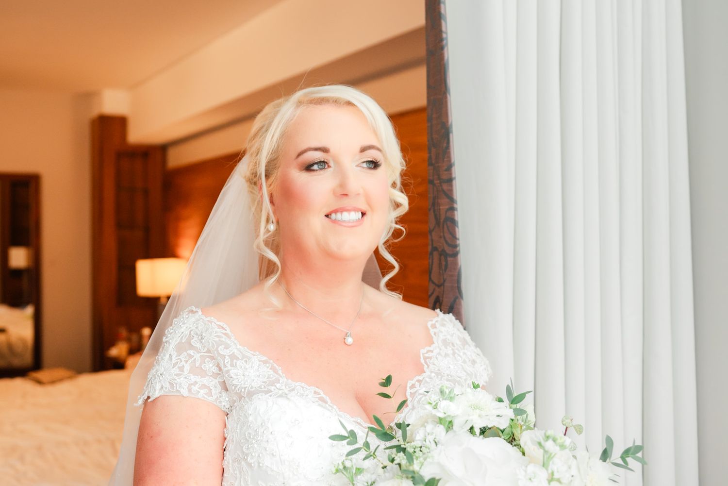 Smiling bride in white lace gown with veil holds a delicate green and white floral bouquet.