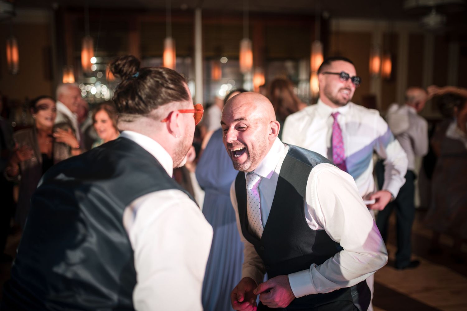 Two men in formalwear laugh and share a joyful moment on the dance floor at an indoor evening celebration.