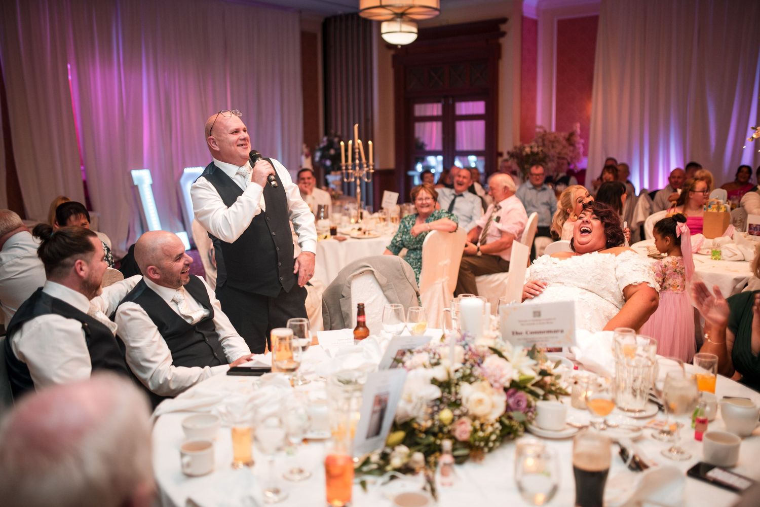 A person in a vest gives a speech at an elegantly decorated wedding reception with tables of guests and pink lighting.