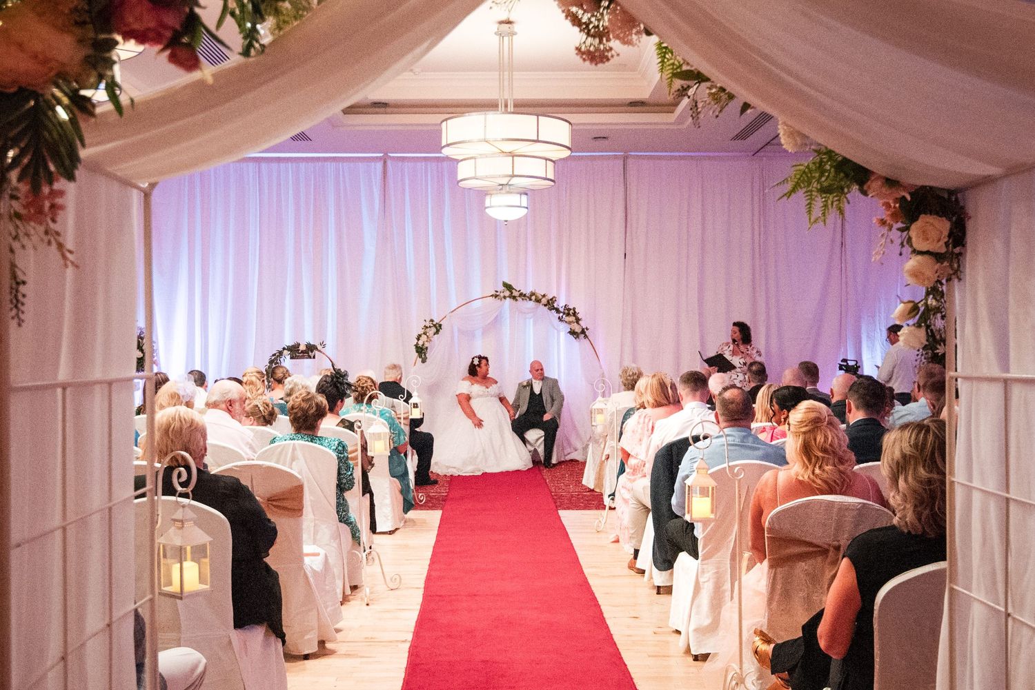 Wedding ceremony venue with draped white fabric, chandeliers, floral arches and red carpet aisle in elegant indoor setting.