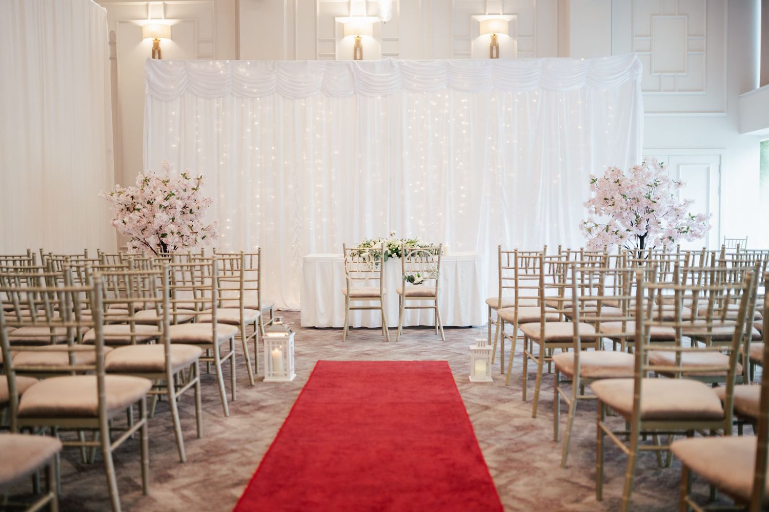 An elegant white wedding ceremony space with red carpet aisle runner and gold chairs decorated with hanging star lights.