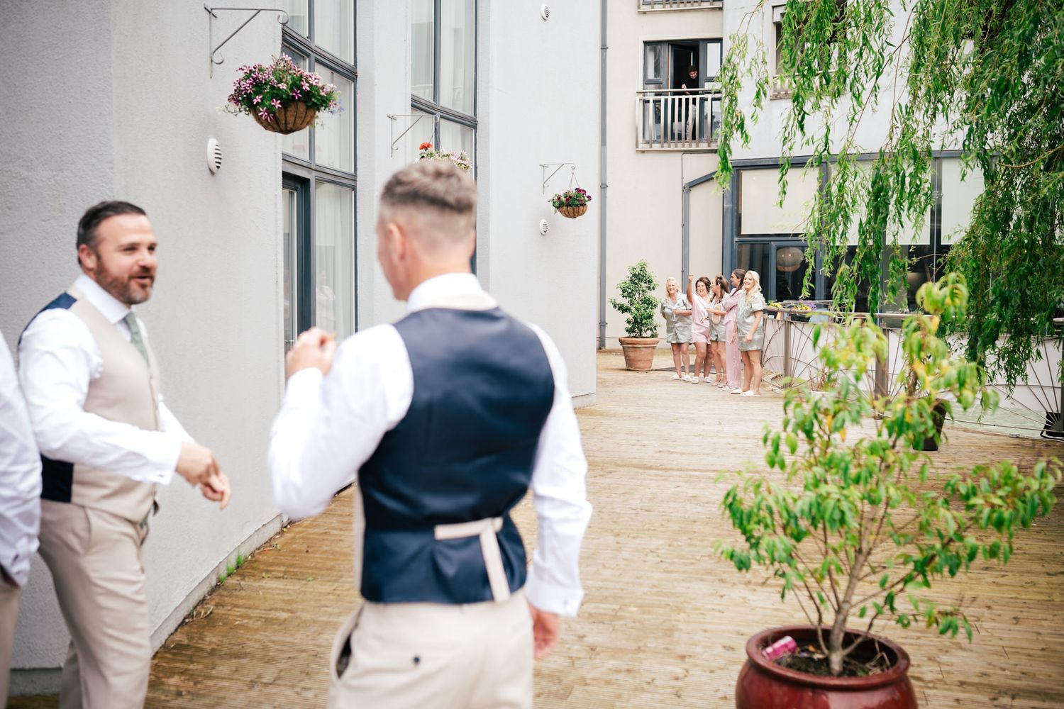 Two people dressed in wedding attire chat while standing in a courtyard with a weeping willow tree and potted plants.