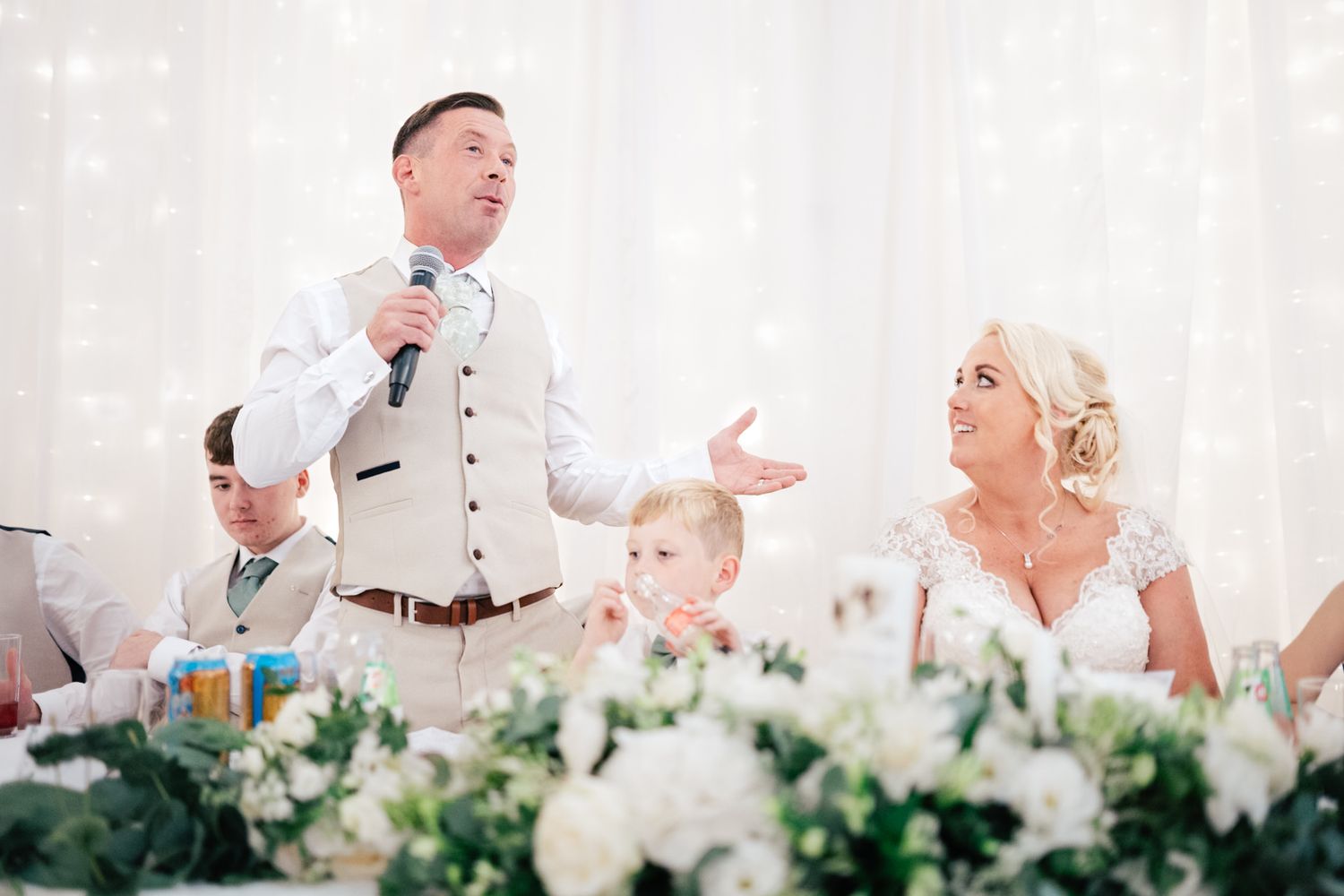 Reception speech moment at elegant white wedding with floral centerpieces and table decorations.