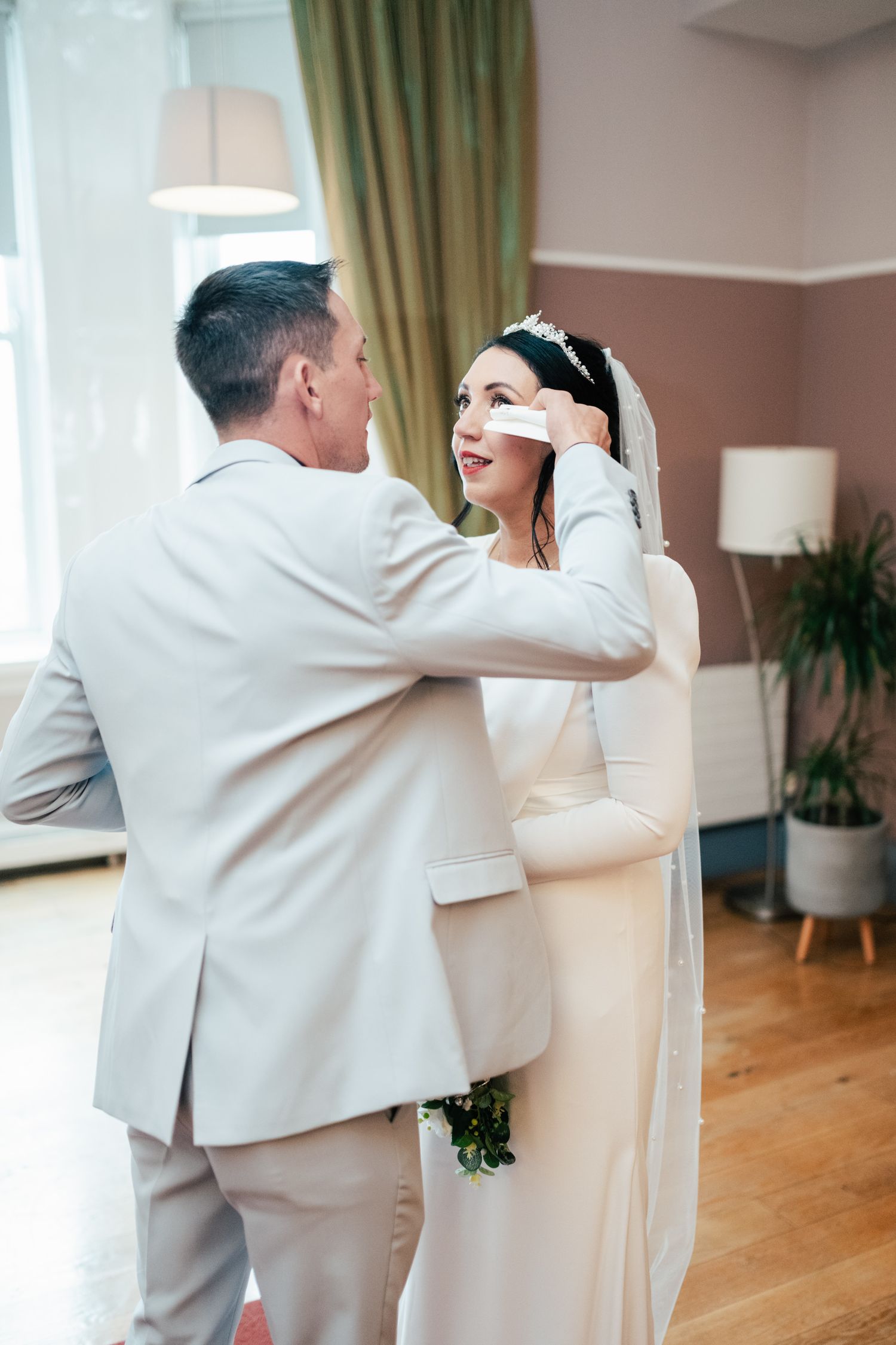 Two people share a tender moment in white attire at an indoor wedding ceremony with green curtains and wooden floors.