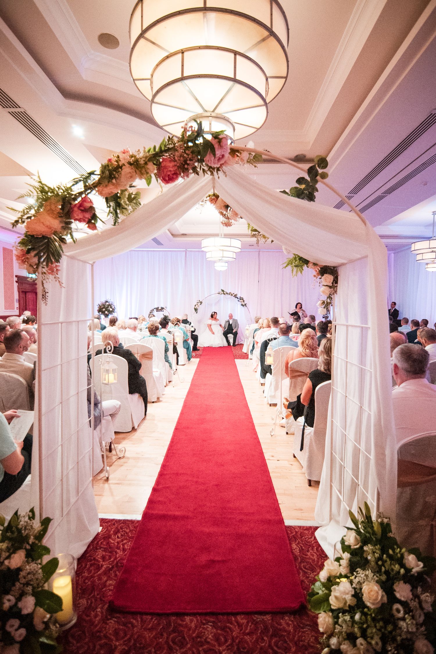 Elegant wedding ceremony aisle with red carpet, white fabric arch adorned with floral arrangements, and crystal chandeliers above.
