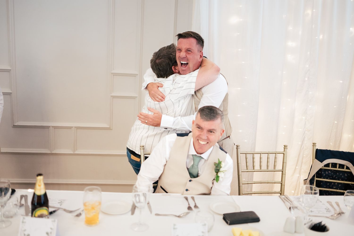 Wedding guests share a joyful moment together at a reception table decorated with white linens and gold chairs.