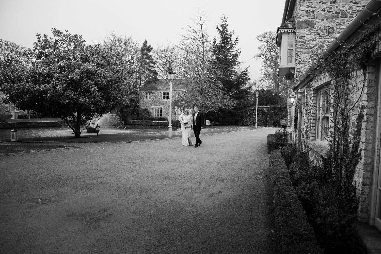 A black and white photograph shows two figures walking on a path between stone buildings in a rural setting.