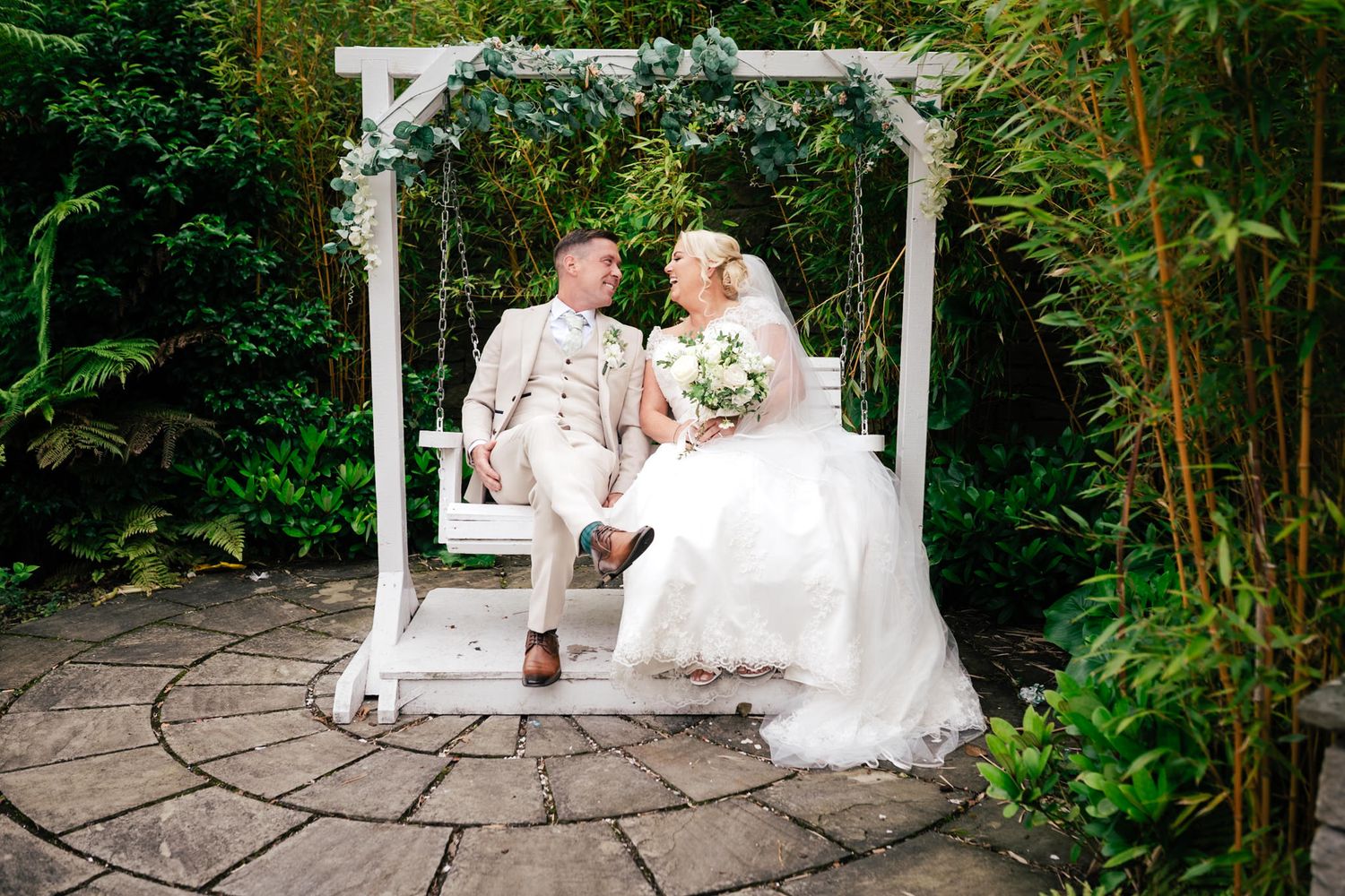 A couple shares a moment under a white garden arbor surrounded by lush greenery and bamboo plants.