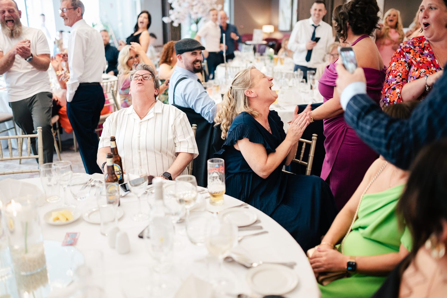 Candid shot of wedding guests laughing and enjoying themselves at a reception with round tables and festive decor.