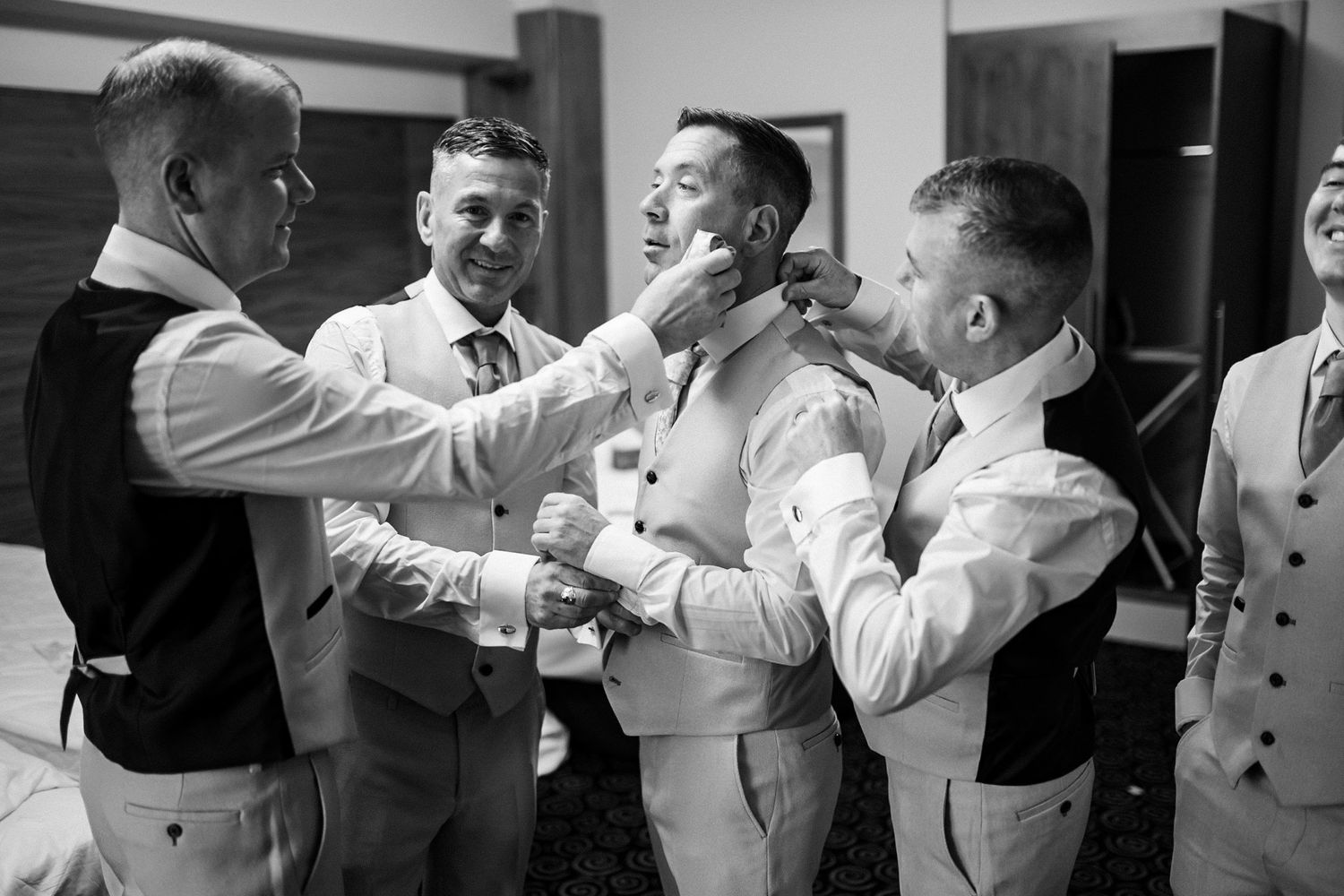 Groomsmen helping each other get ready by adjusting bow ties and boutonnières in formal wedding attire.
