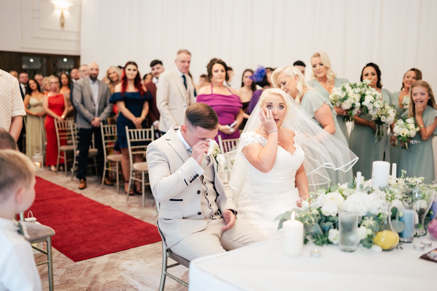 Emotional reaction during a wedding ceremony with guests seated in a reception hall adorned with a red aisle runner.