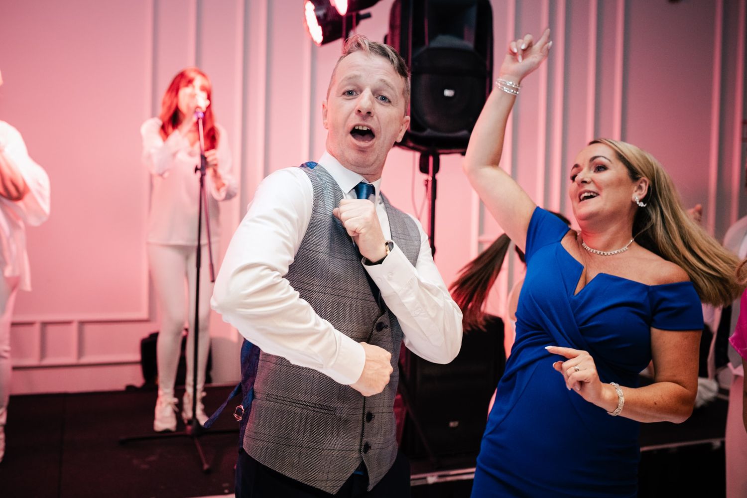 Two energetic wedding party guests dance enthusiastically on the dance floor at a reception with pink walls.