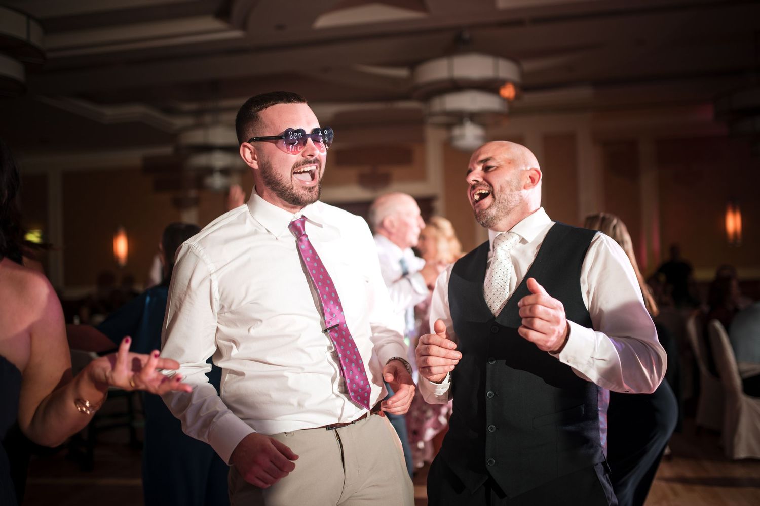 Groomsmen celebrate on the dance floor at a dimly lit wedding reception.