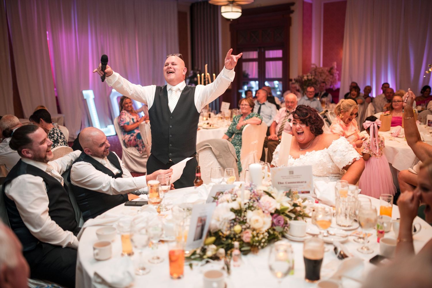 An energetic speaker raises their arms while delivering a toast at a formal wedding reception surrounded by tables of guests.