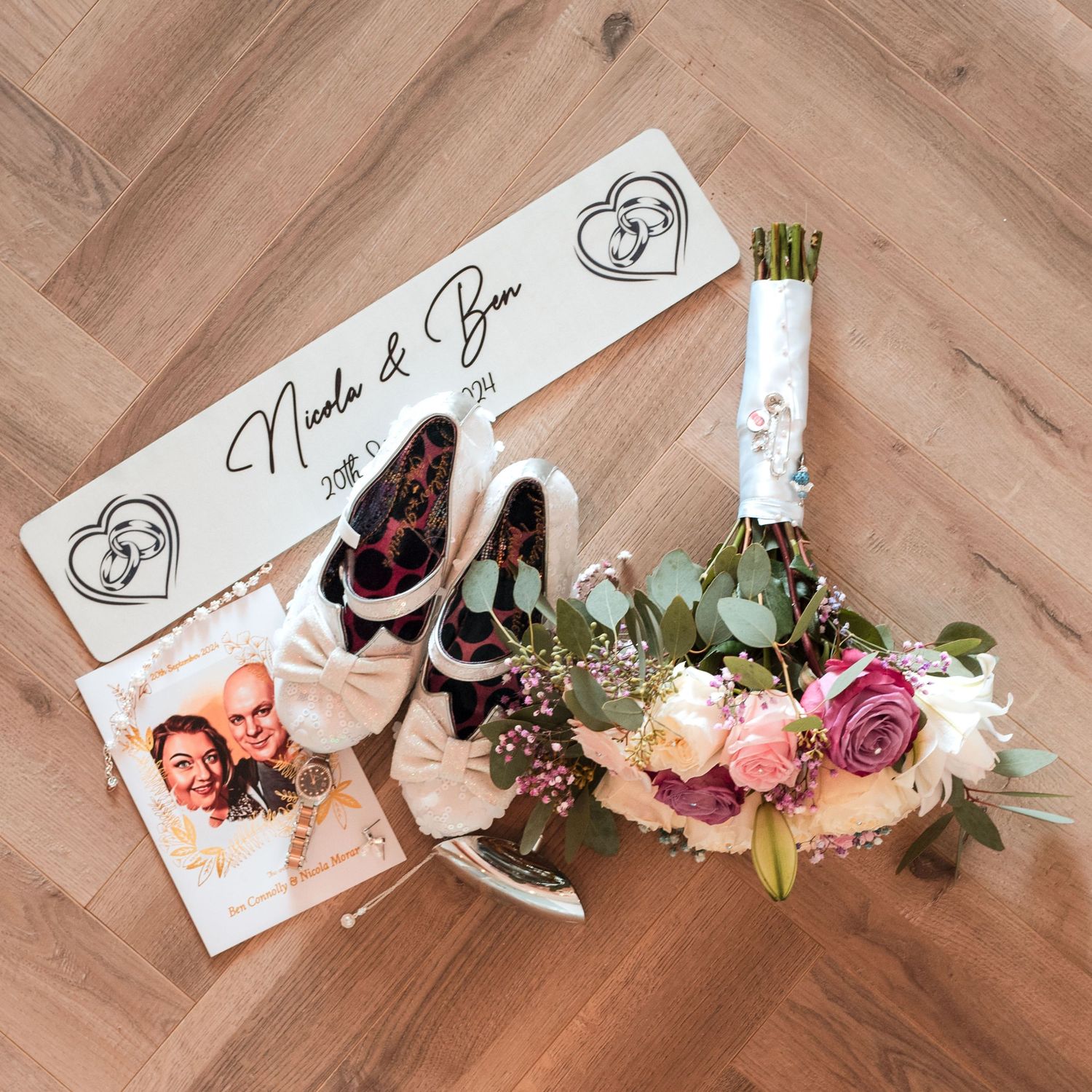 Wedding flat lay with bridal shoes, bouquet of pastel roses and eucalyptus, and a personalized name sign on wooden flooring.