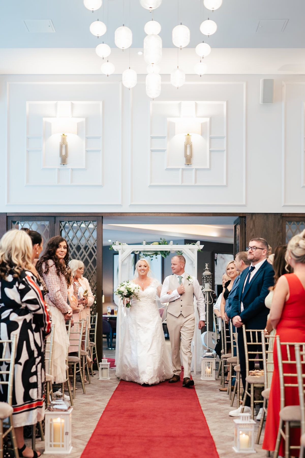 Wedding ceremony in progress with couple walking down red-carpeted aisle while guests look on under elegant lighting.
