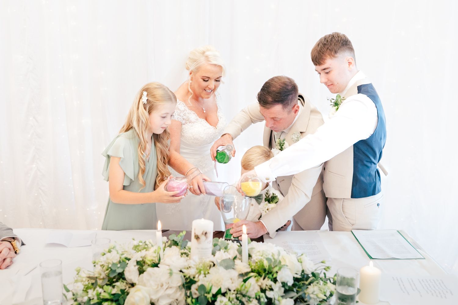 Family pouring colorful sand into glass container during unity ceremony with floral arrangements on white table.