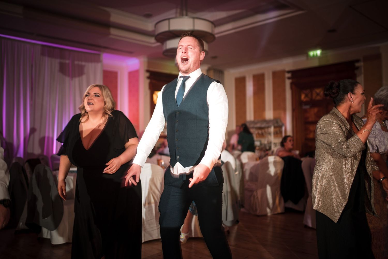 Wedding party guests dancing and celebrating energetically on a dance floor with purple uplighting in a ballroom.