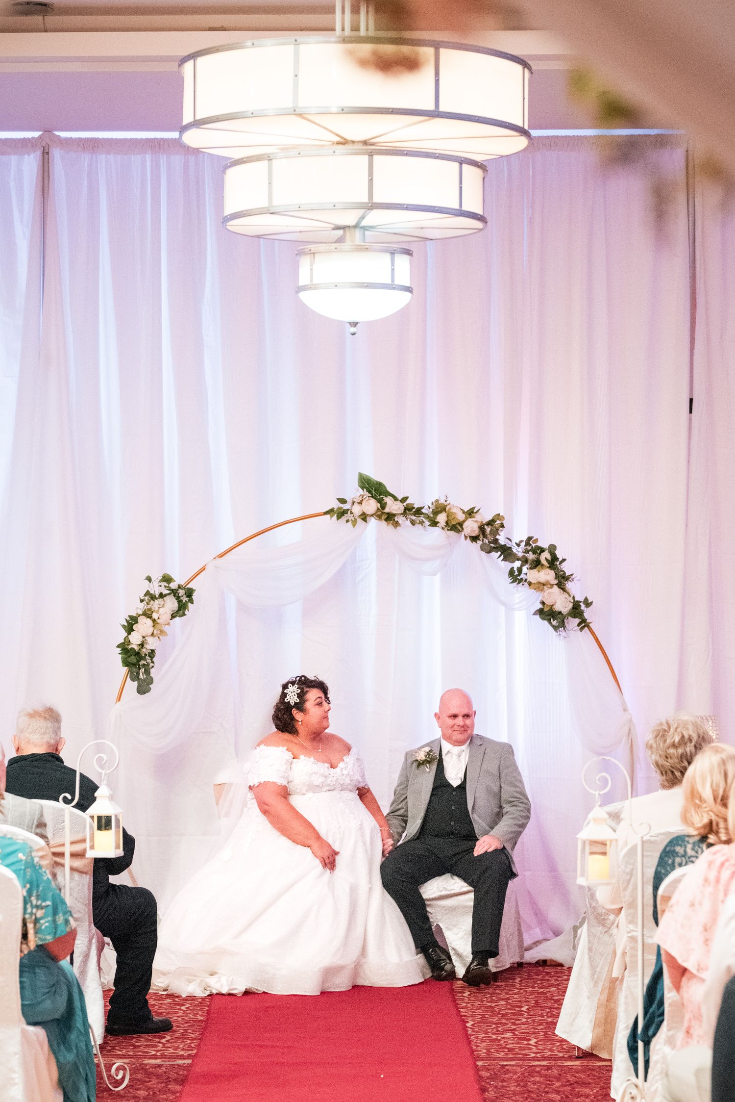 Wedding ceremony scene with floral arch, modern tiered lighting fixtures, and red carpet in an elegant indoor venue.