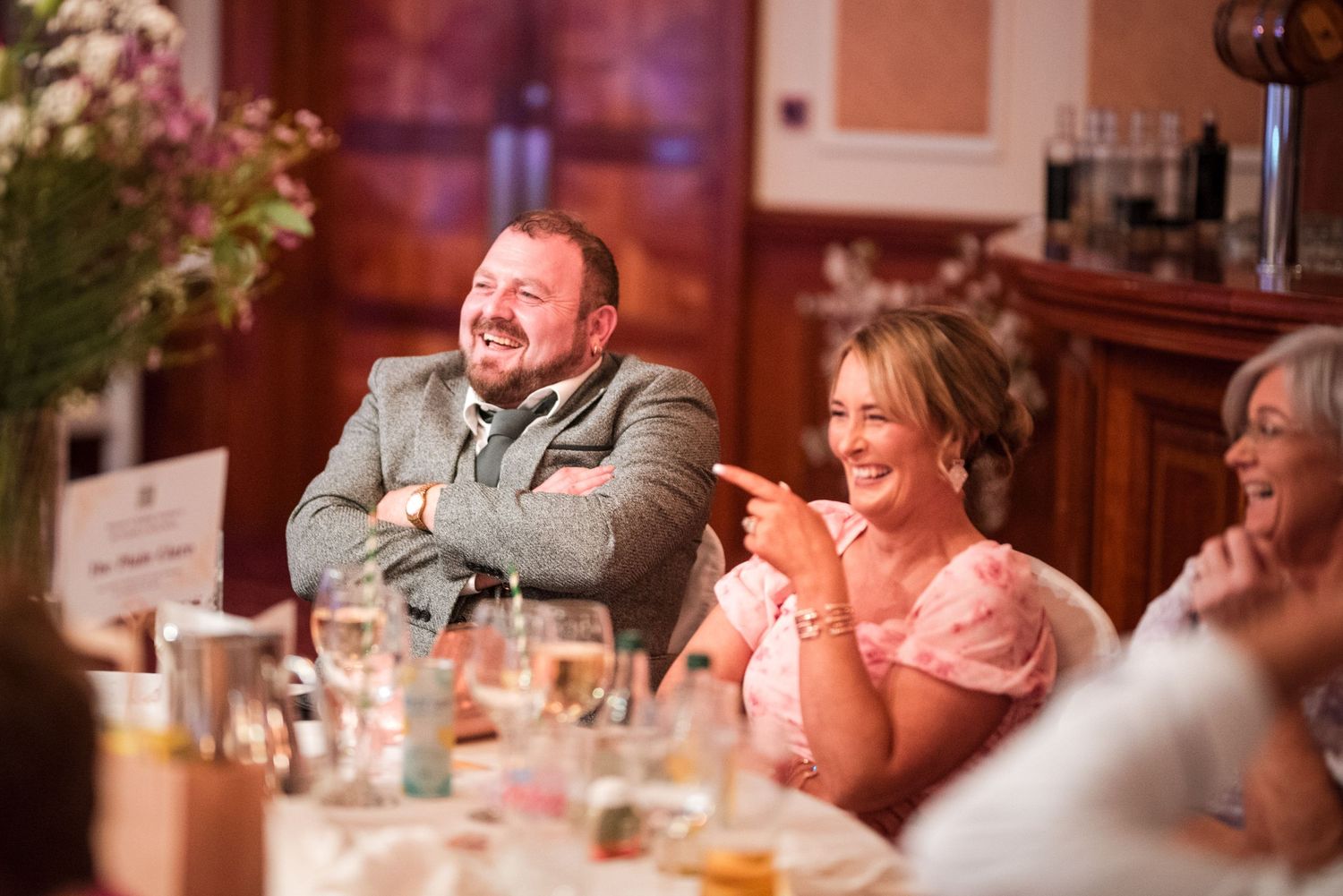 Wedding guests enjoying laughter and conversation at a reception dinner table in warm lighting.