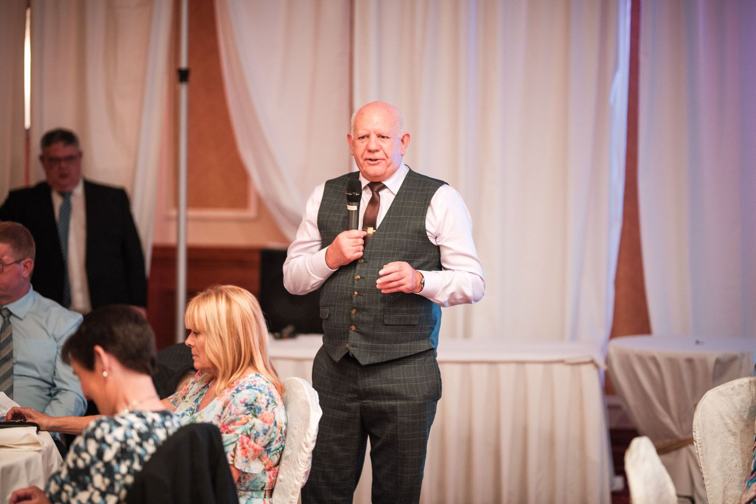 A festive wedding reception shows guests engaging in lively conversation and dancing at an elegant indoor venue.