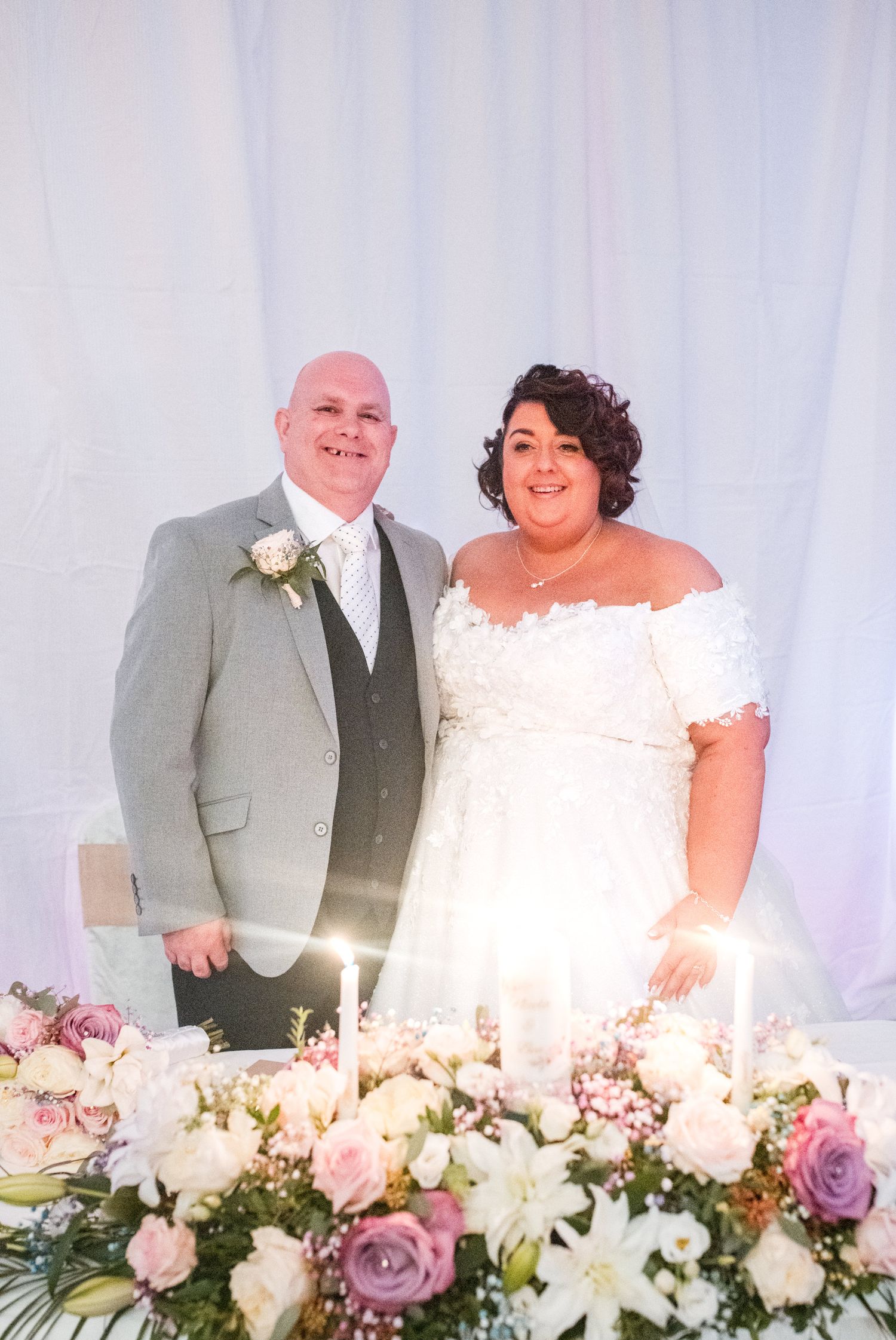 Wedding couple seated at head table with elegant white and purple floral arrangements.