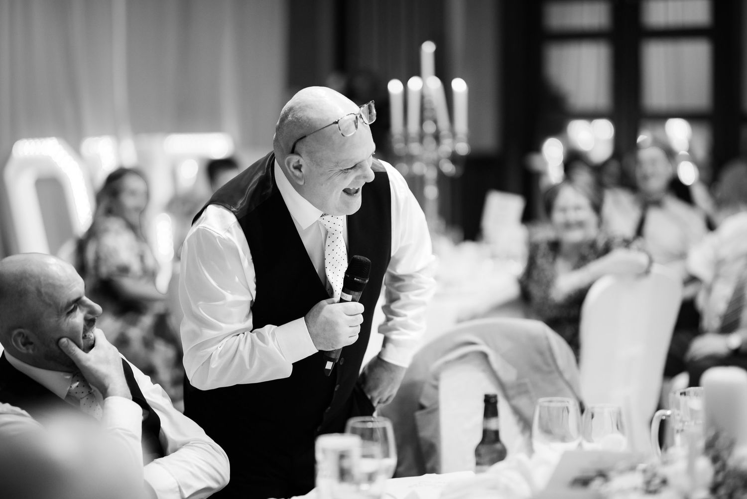 A person giving a speech during a candlelit dinner reception as guests listen attentively in a black and white photo.
